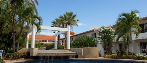 A swimming pool with a waterfall feature, surrounded by palm trees and a white building with a red roof.