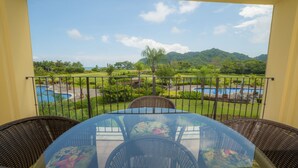 Ein Balkon mit Glastisch und Korbstühlen, mit Blick auf einen Pool und üppiges Grün.