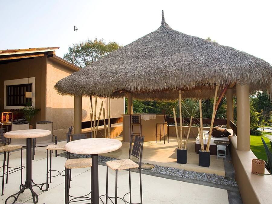 Un espacio al aire libre con techo de paja, con mesas y sillas, rodeado de un jardín.