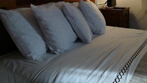 Una cama bien hecha con sábanas y almohadas blancas, un cabecero de madera, una lámpara de noche y un cuadro abstracto y colorido en la pared.