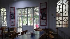 Un café con una mesa central rodeada de sillas, ventanas amplias con rejas decorativas y carteles en la pared.