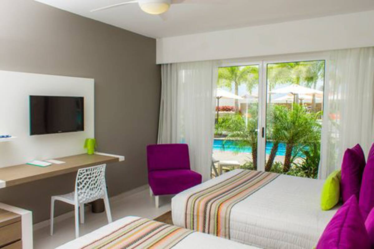 A hotel room with a bed, a desk, a chair, a TV, and a view of a pool and greenery.
