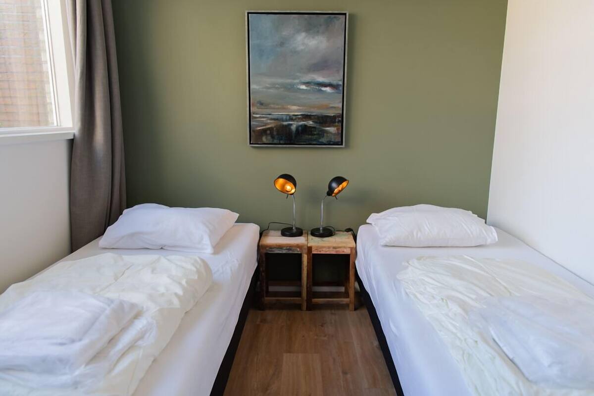 A room with two single beds, a wooden nightstand with two lamps, a painting on the wall, and a window with curtains.