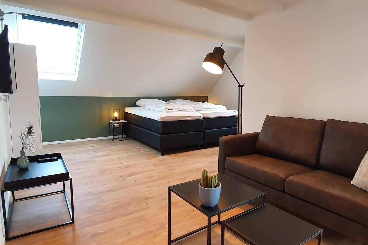A modern living room with a sofa, a coffee table, a TV, a bed with a headboard, a floor lamp, and a small table with a plant.