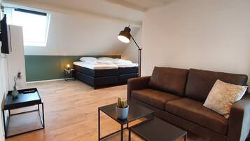 A modern living room with a sofa, a coffee table, a TV, a bed with a headboard, a floor lamp, and a small table with a plant.