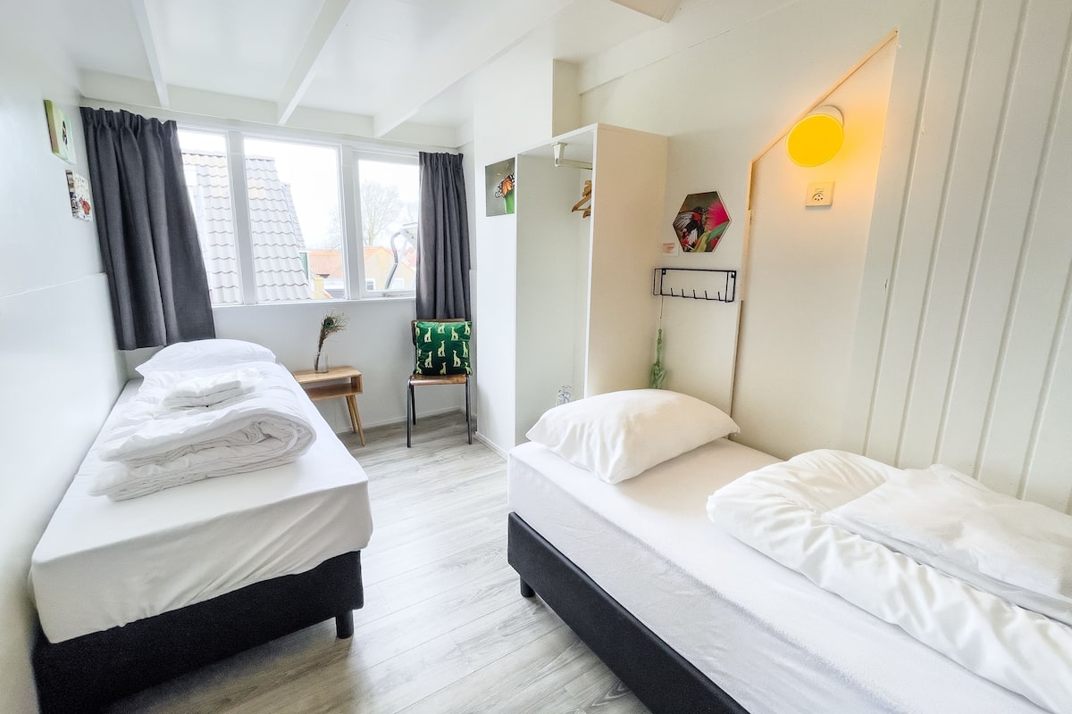 A room with two beds, a window with curtains, a small table with a plant, and a wall-mounted light fixture.