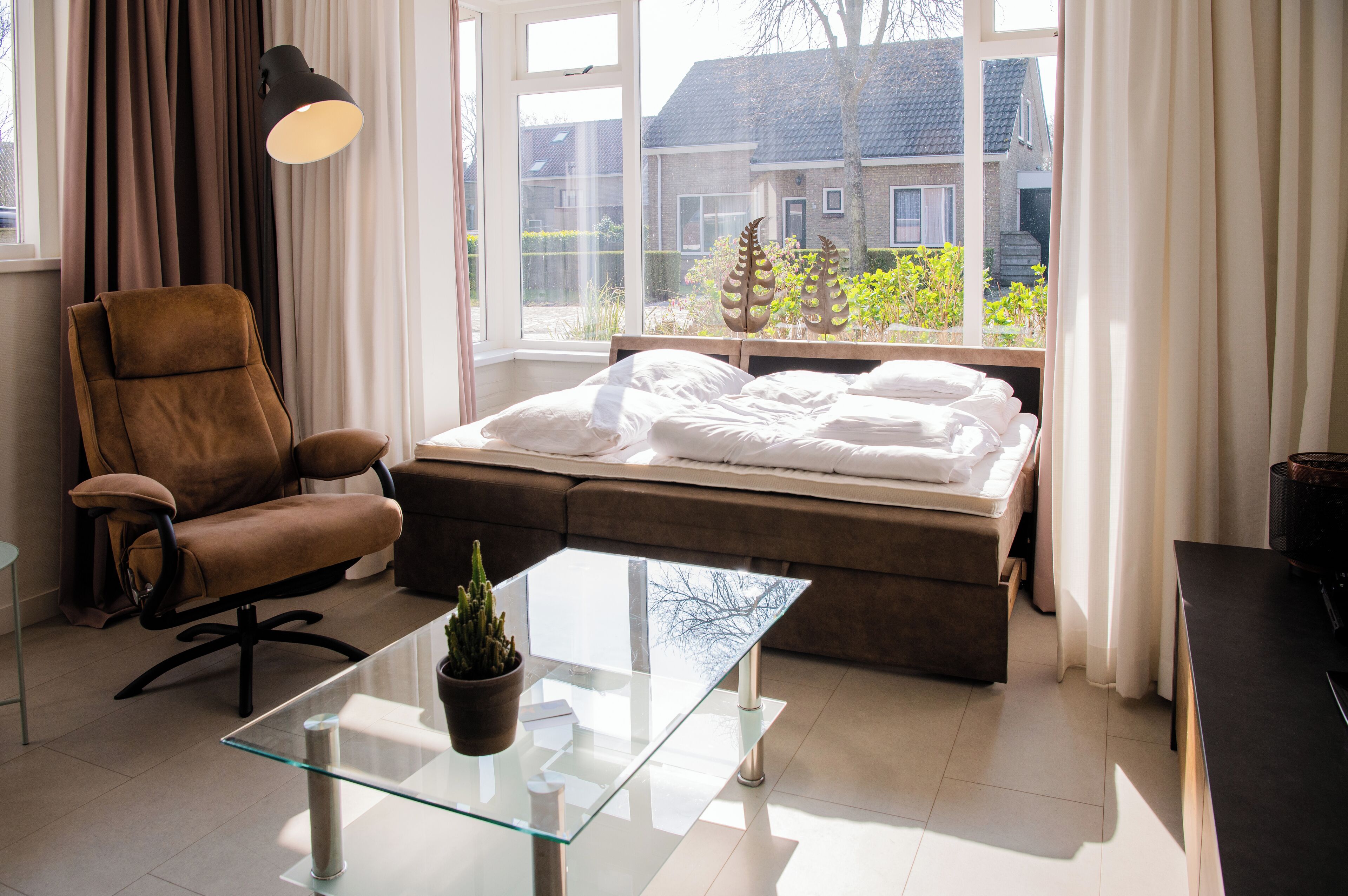 A modern bedroom with a bed, a chair, a glass table, and a view of a house through a window.