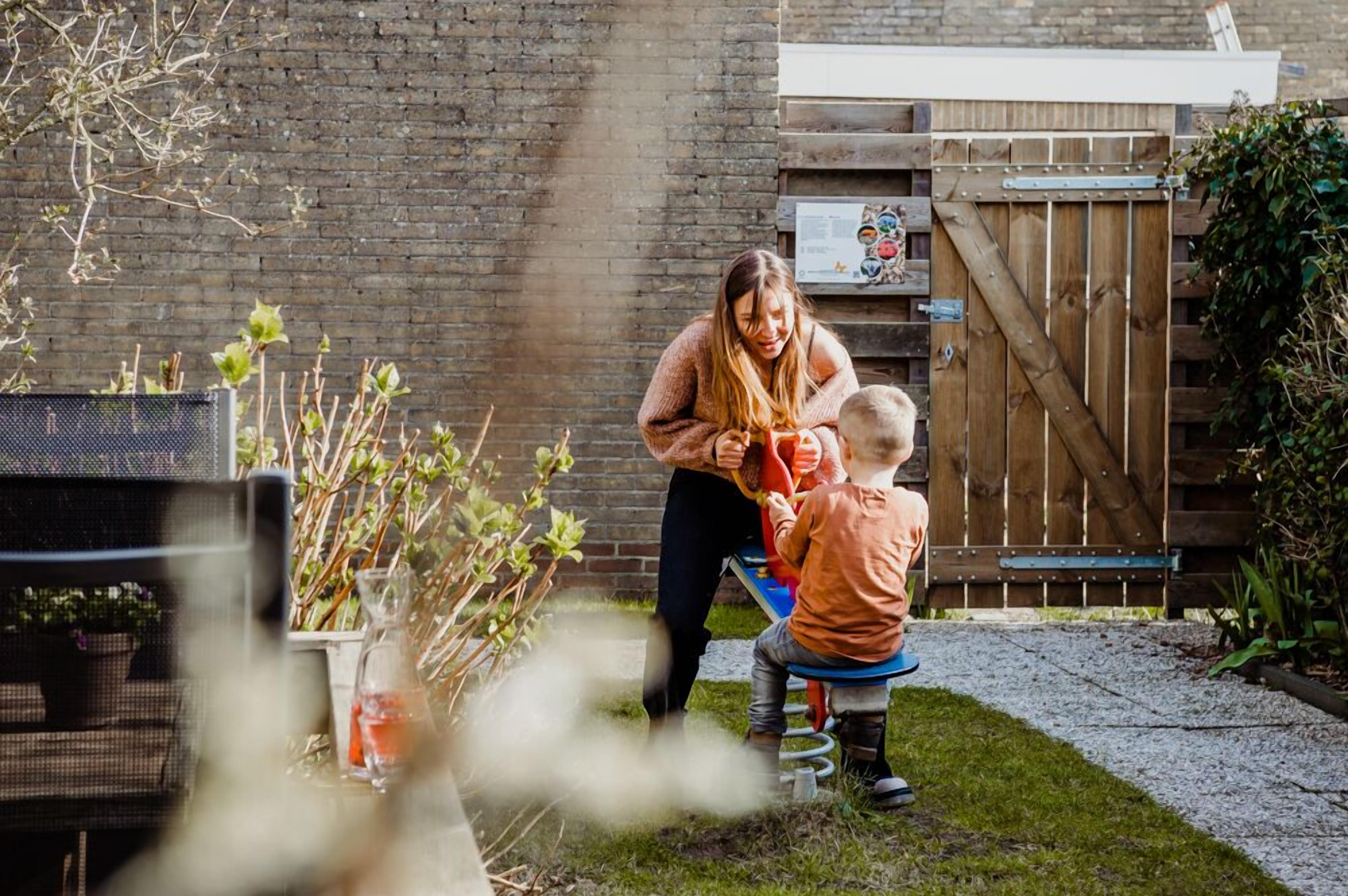 Eine Frau und ein Kind spielen mit einem Spielzeug in einem Garten mit Holzzaun, Backsteinmauer und Blumenbeeten.