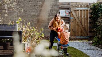 Eine Frau und ein Kind spielen mit einem Spielzeug in einem Garten mit Holzzaun, Backsteinmauer und Blumenbeeten.