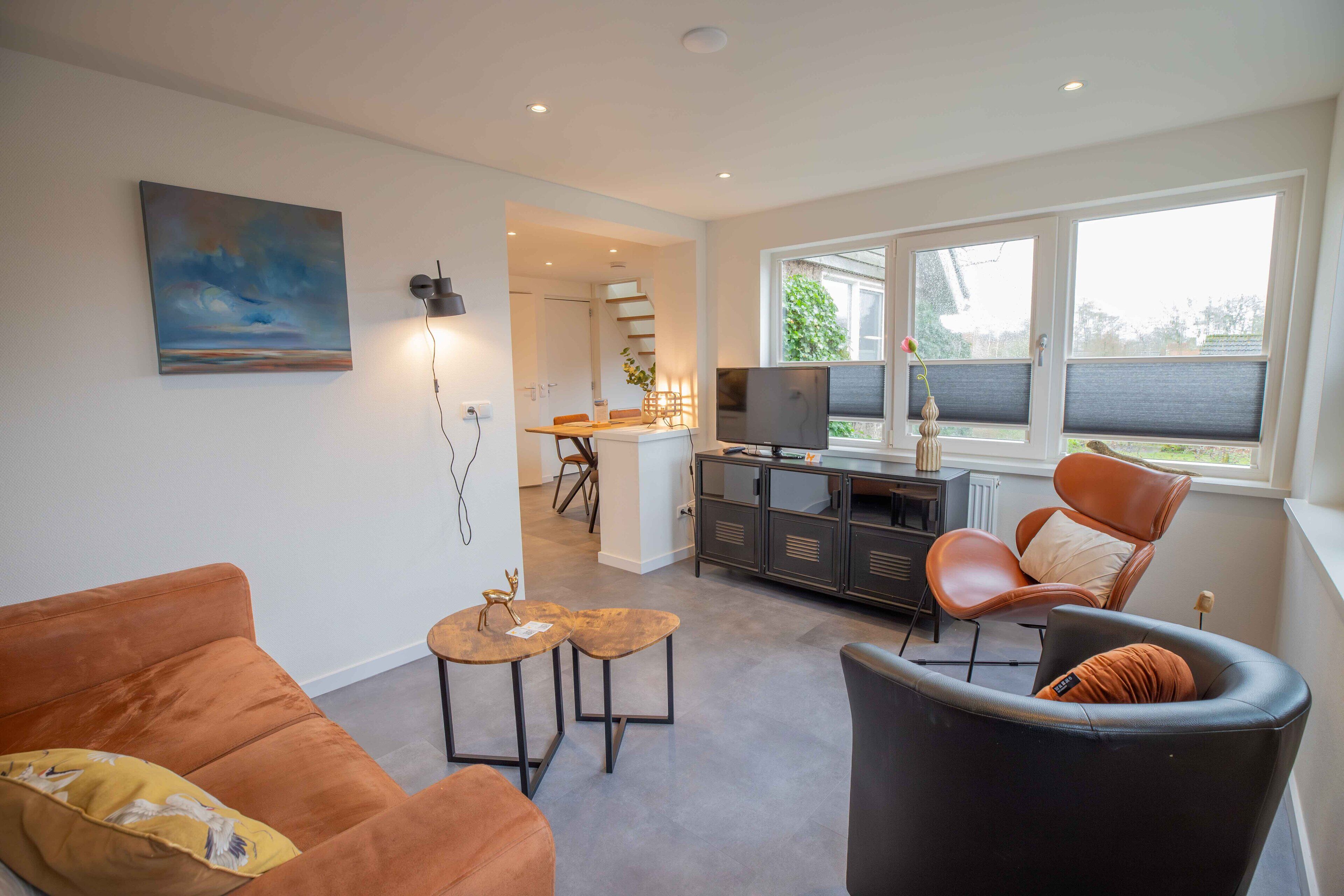 Un salon moderne comprenant un canapé, un fauteuil, une petite table et un téléviseur. On y trouve également un coin cuisine avec une table et des chaises, ainsi qu’un escalier menant à un autre niveau.