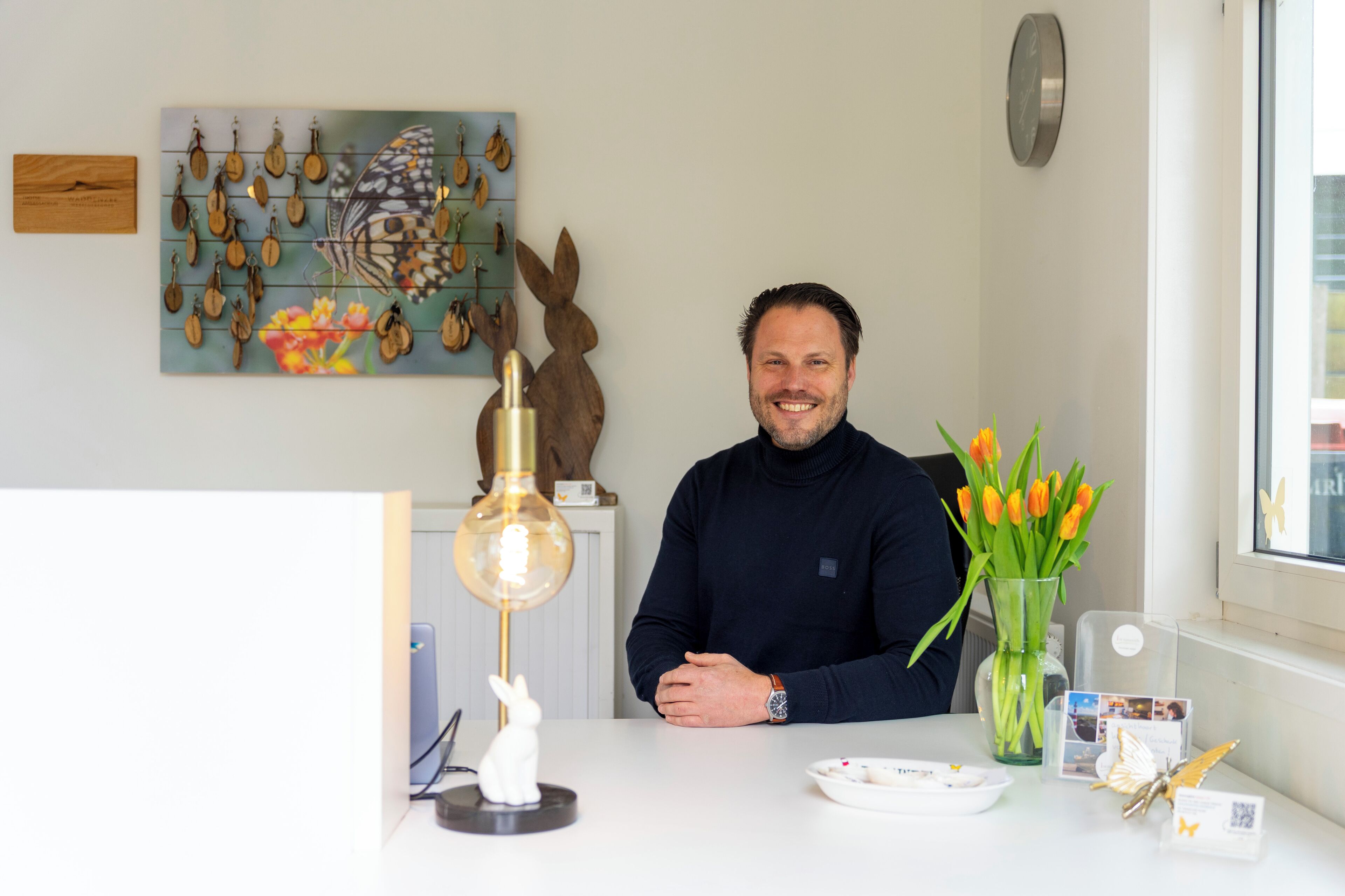 Un homme est assis à une table devant un vase de tulipes, un réveil est accroché au mur et une décoration en forme de papillon est posée à proximité.