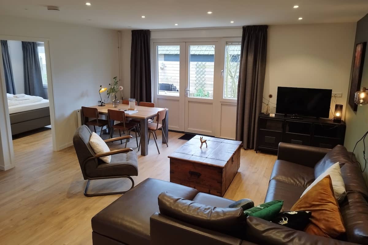 A modern living room with a sofa, a dining table with chairs, a TV stand, and a bed in the background.