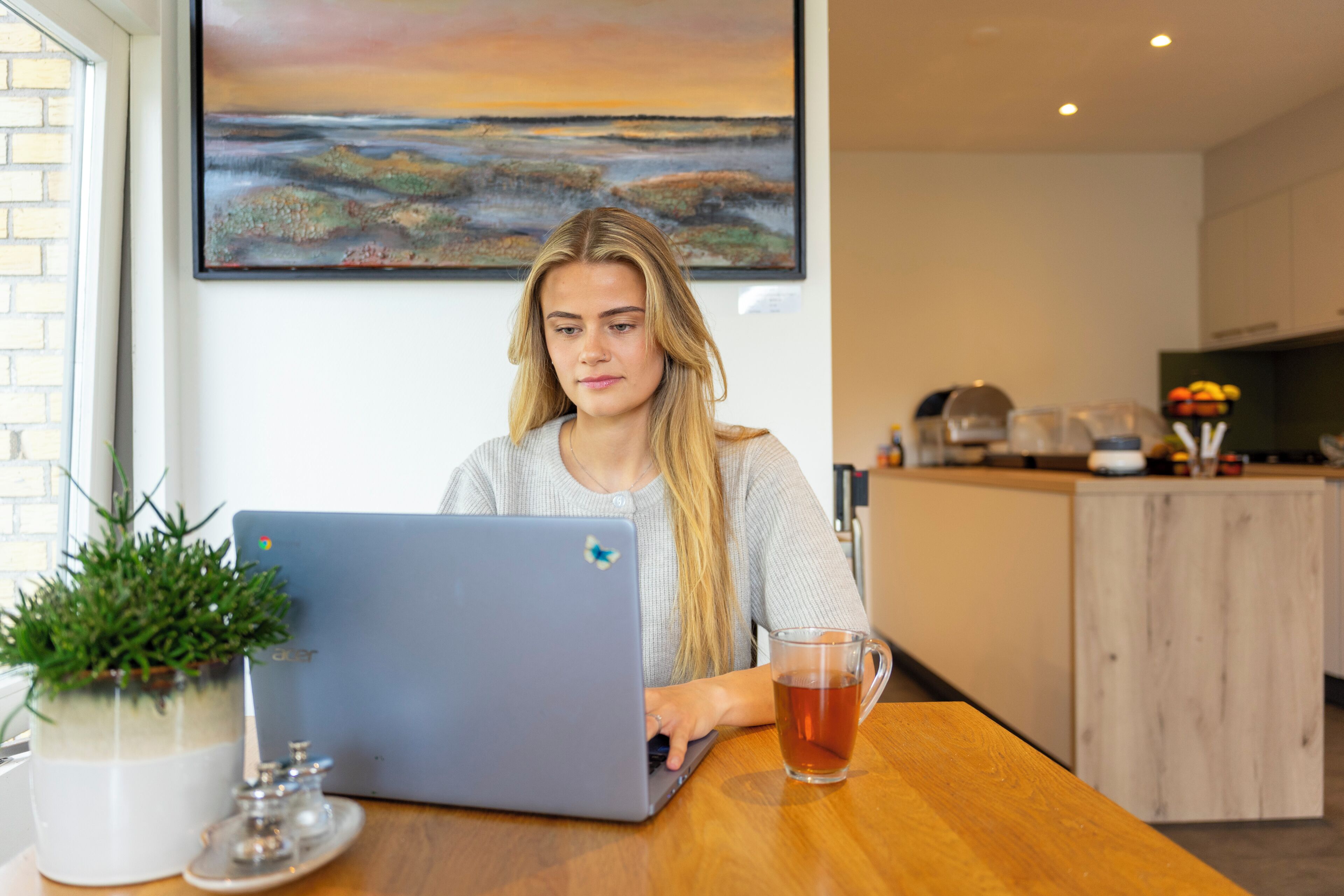 A person using a laptop at a wooden table with a potted plant and a glass mug.