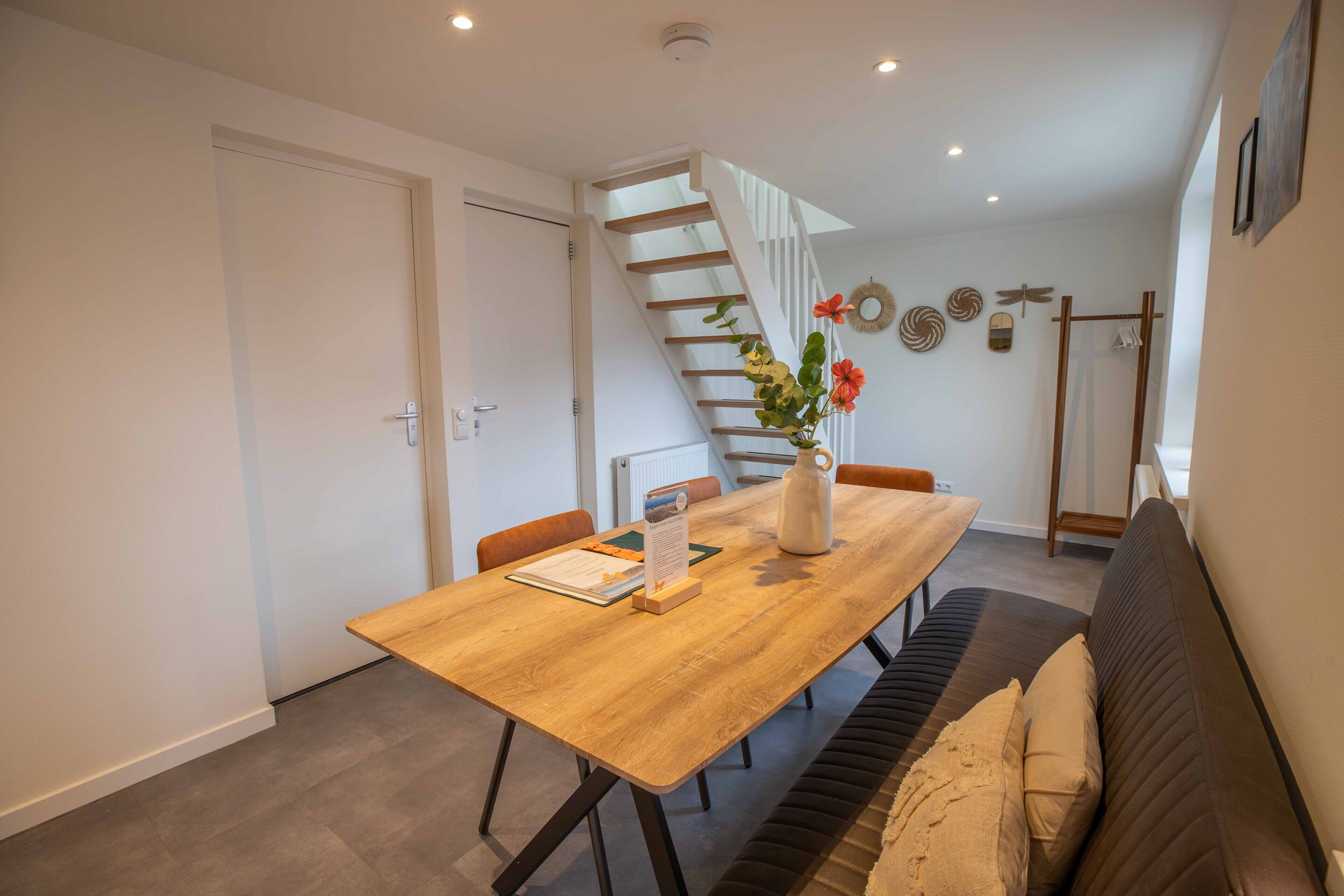 A dining area with a wooden table, chairs, a vase with flowers, a staircase, and a coat rack.