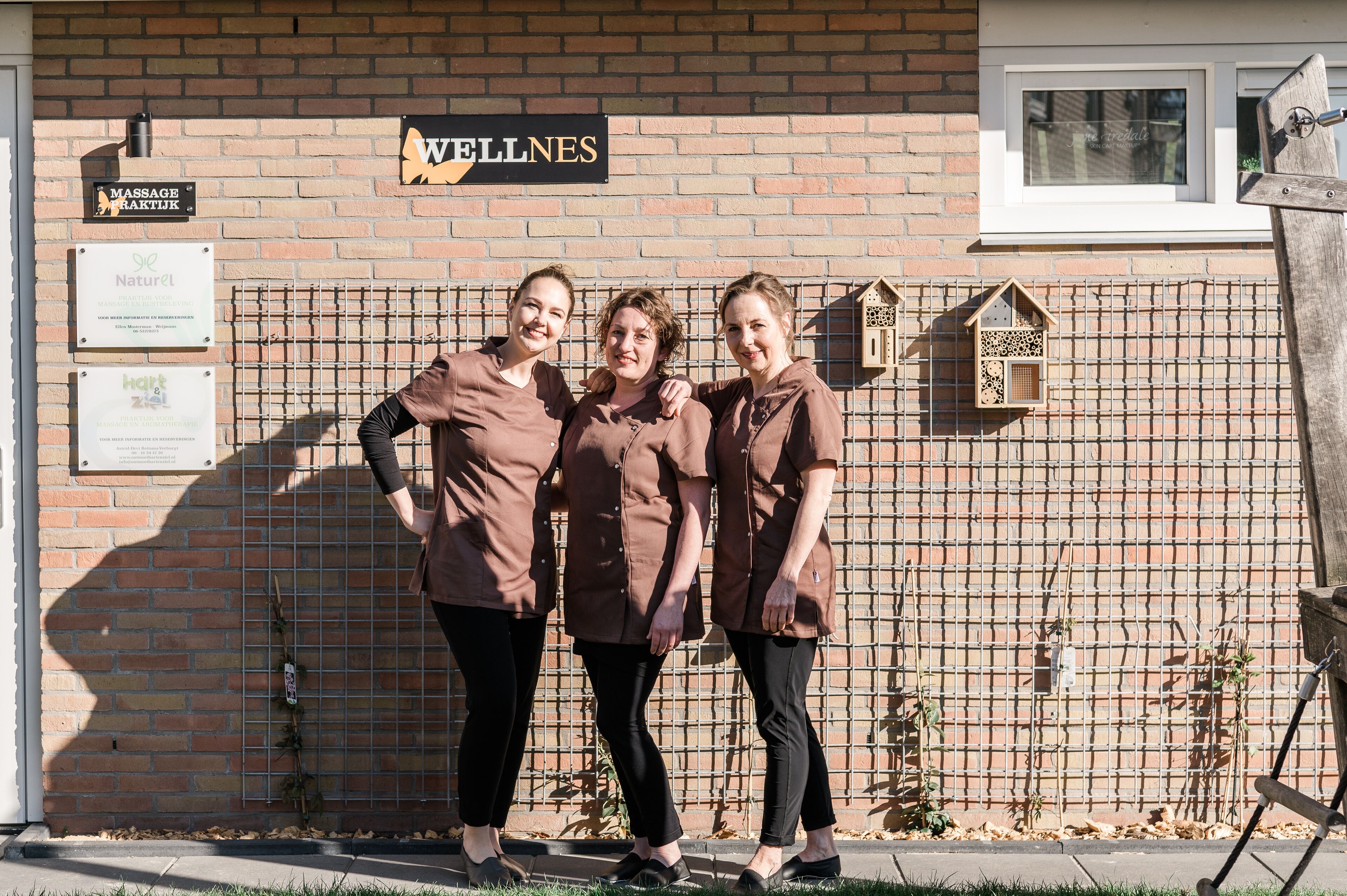 Three individuals standing in front of a brick wall with a sign that reads "WELLNES."