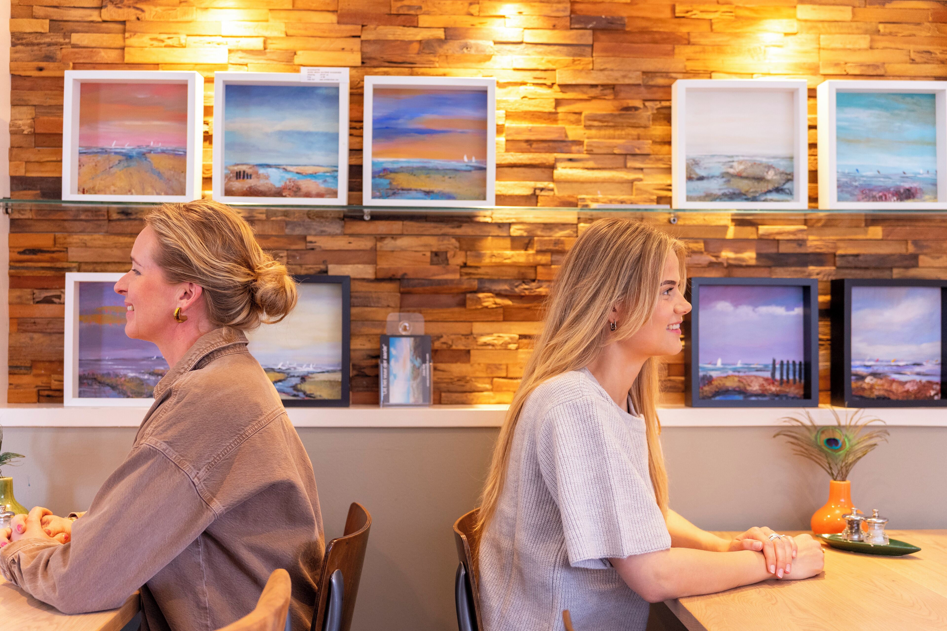 Dos personas sentadas en una mesa, con una pared de madera y obras de arte enmarcadas al fondo.