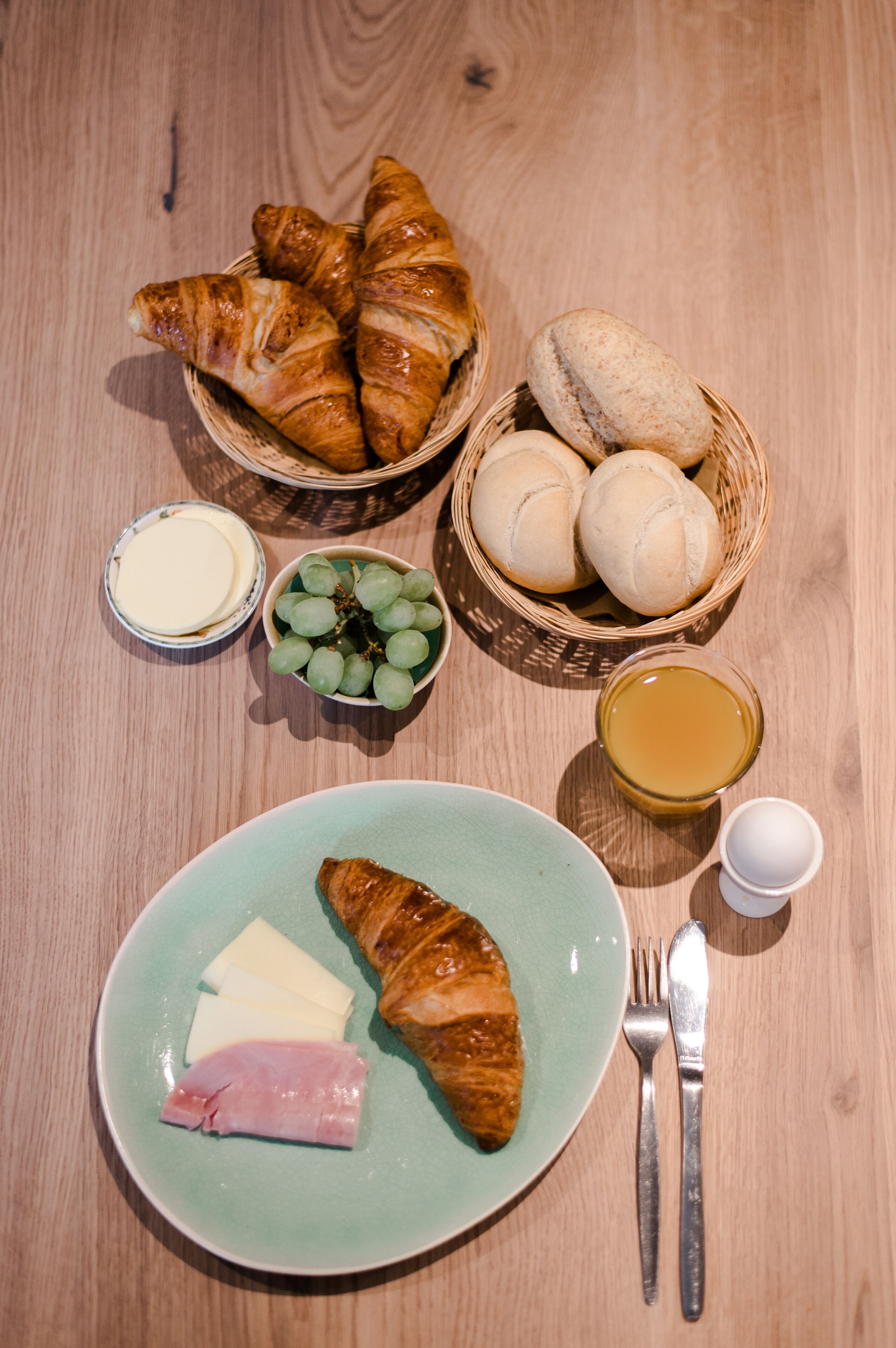 A wooden table with a plate of food, including croissants, cheese, and ham, along with a bowl of grapes and a small container of butter.