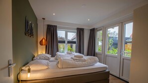 A modern bedroom with a large bed, a bedside lamp, and a view of the outside through large windows. - De Vlindertuin Ameland (Nes)