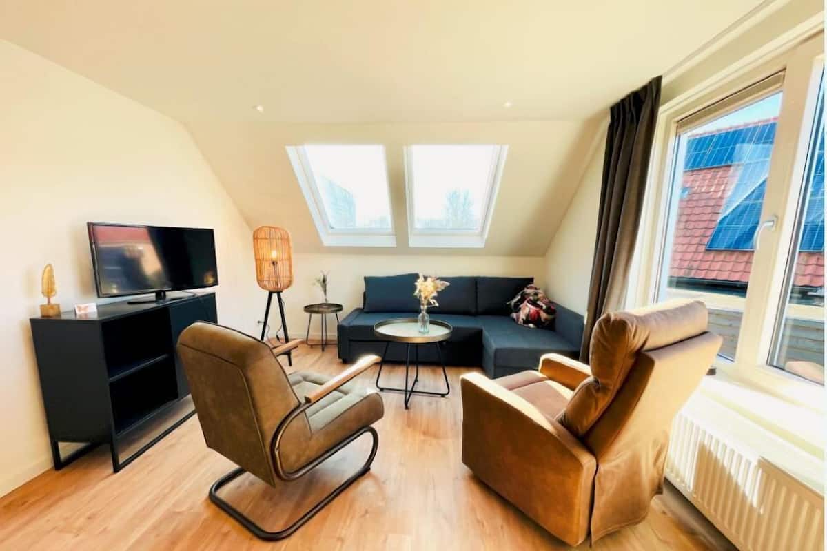 A modern living room with a flat-screen TV, two sofas, a coffee table, and a floor lamp. There are skylights above the sofas and a large window on the right.