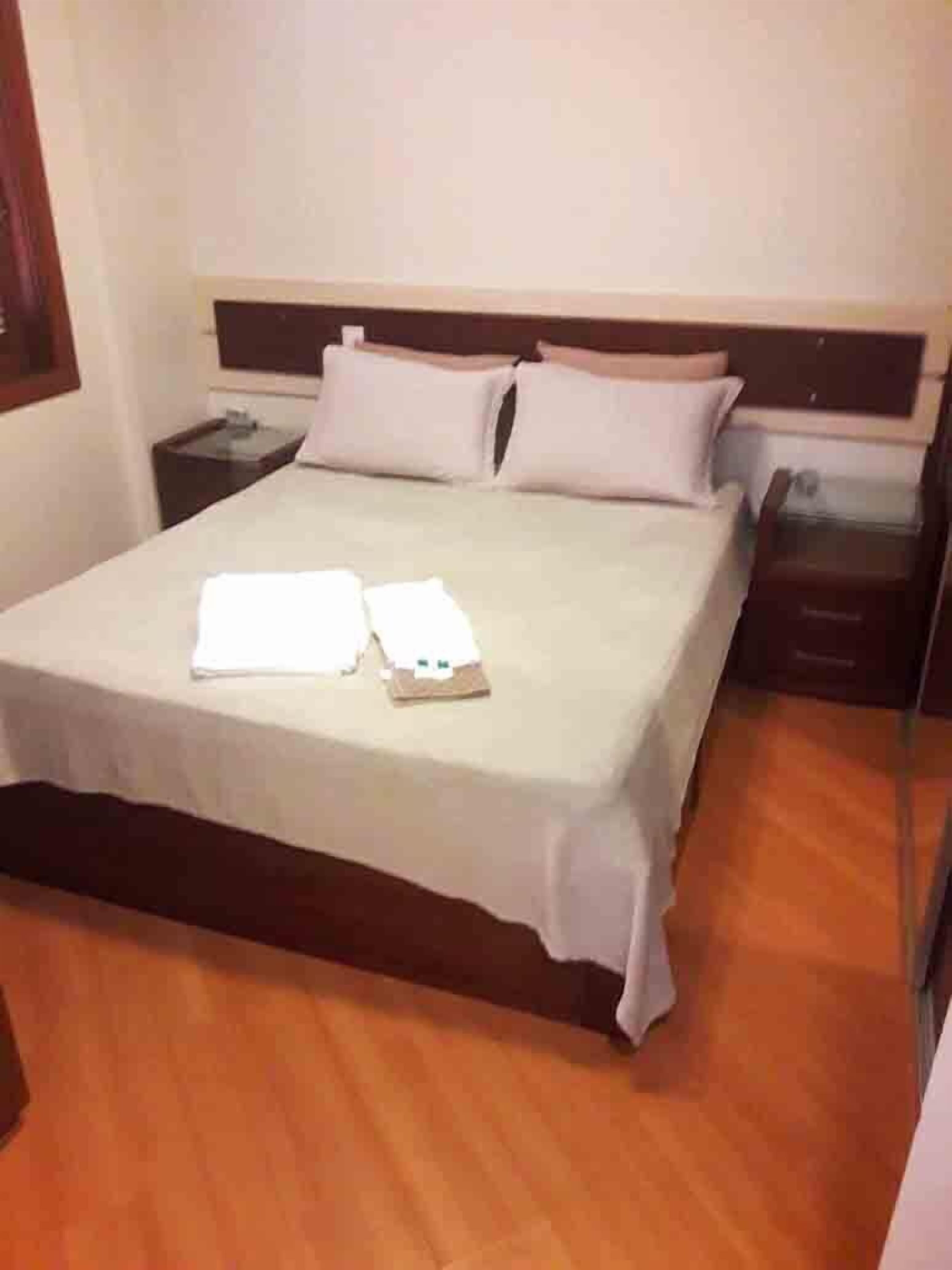 A neatly made bed with a white bedspread and two pillows, a bedside table with a lamp, and a wooden floor.