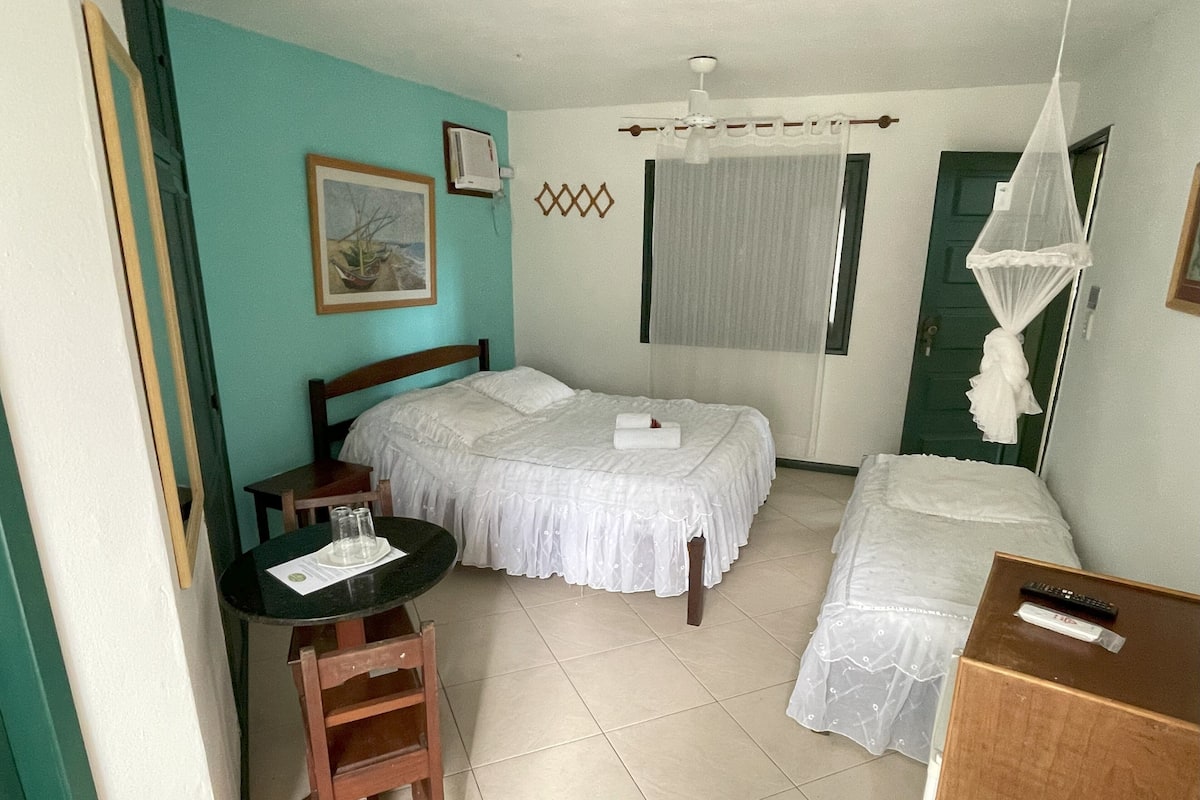 A room with two beds, a small table with glasses, a chair, a green door, and a painting on the wall.