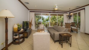 Un salón con sofá, televisión, mesita de madera y lámpara. Se ven palmeras a través de ventanas amplias.