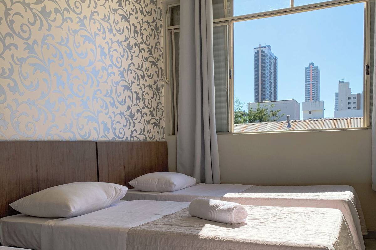 Habitación de hotel con tres camas, papel tapiz estampado, vista a edificios de la ciudad y una ventana con cortinas.