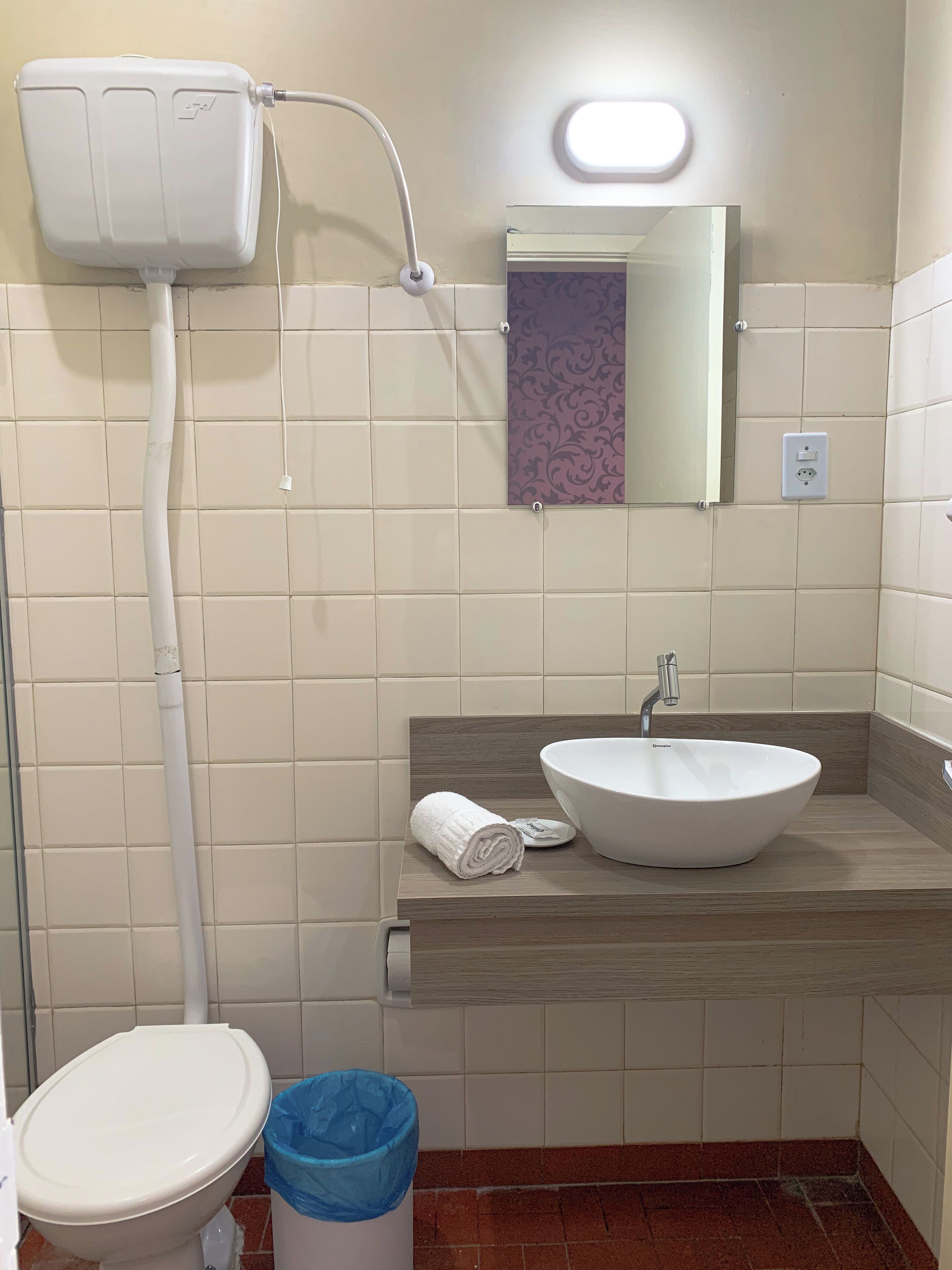 A bathroom with a toilet, a sink with a basin, a towel, a trash bin, and a toilet paper holder.