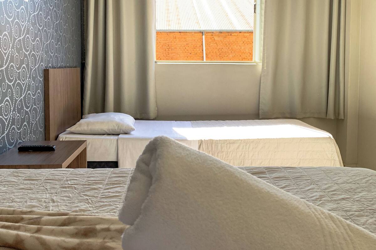 Una habitación de hotel con dos camas, una ventana con cortinas, un papel tapiz estampado y un control remoto sobre una mesita de noche.