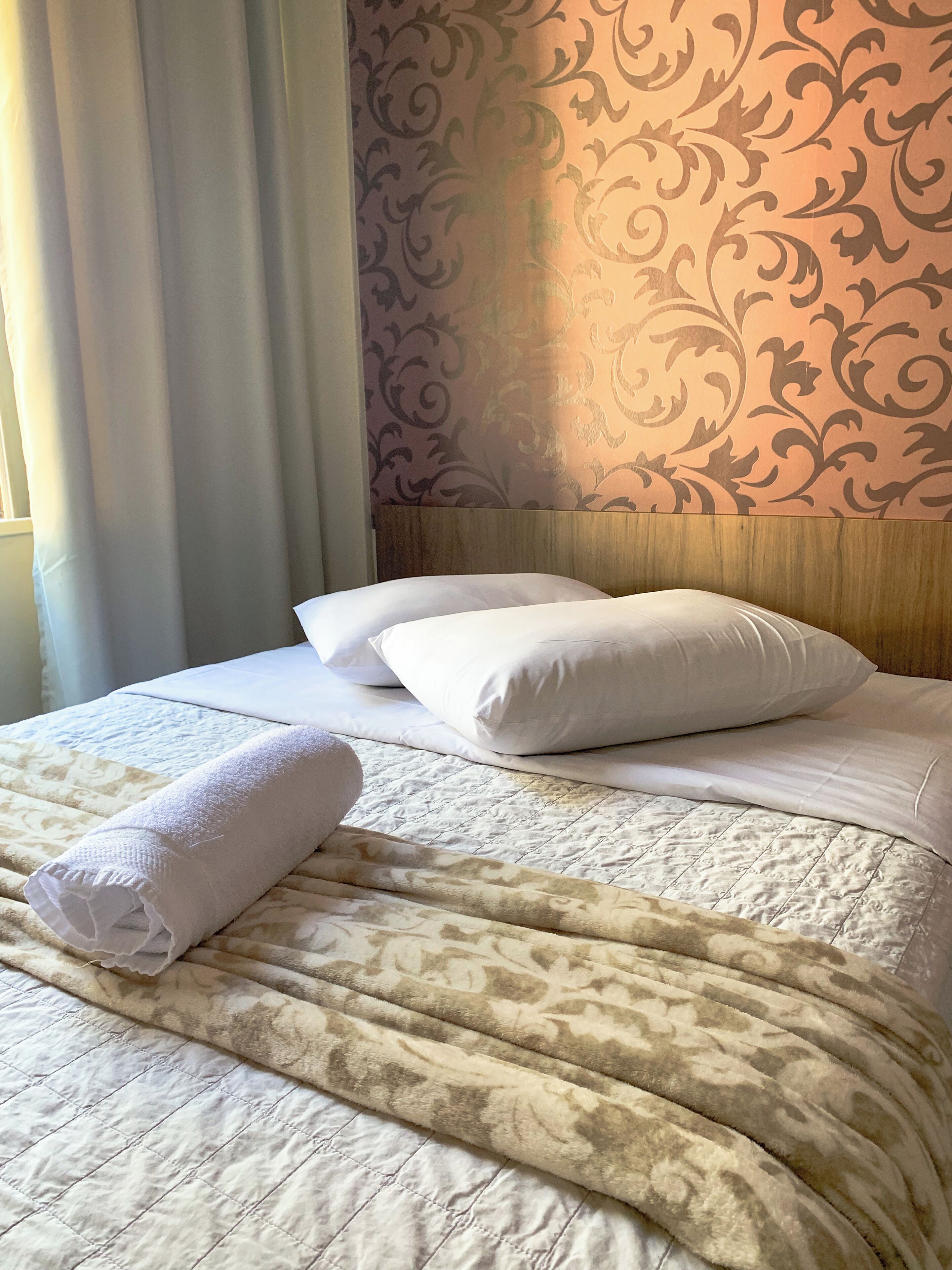 A neatly made bed with white linens, a beige patterned blanket, and a rolled white towel.