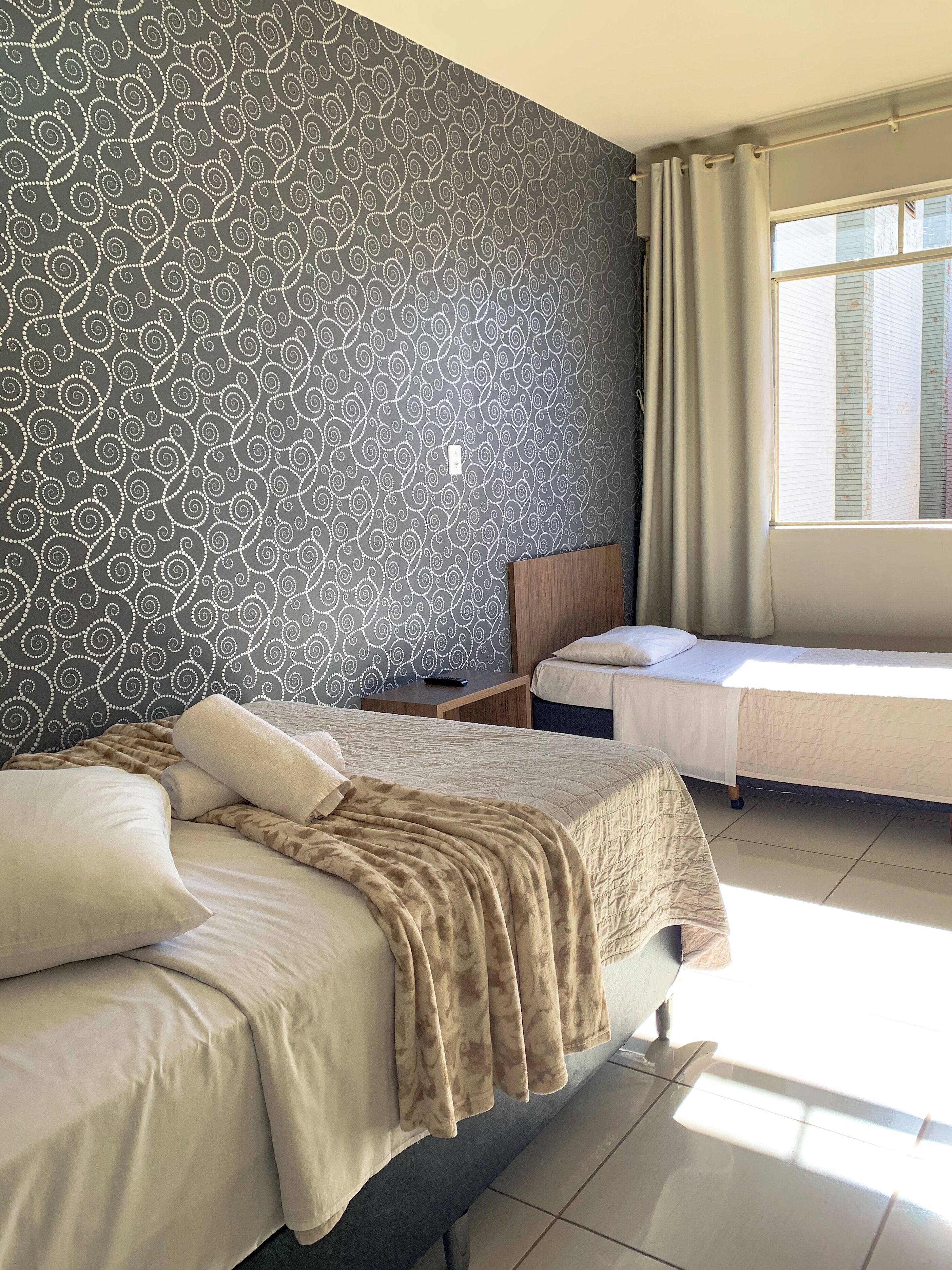 Habitación de hotel con papel tapiz estampado, una cama con manta color beige, una mesita de noche y una ventana con cortinas.
