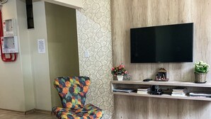Een moderne woonkamer met een houten vloer, een kleurrijke fauteuil, een klein voetenbankje, een flatscreen televisie bevestigd aan een houten muur, en een tafeltje met decoratieve objecten.