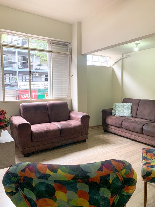 A living room with two sofas, a colorful armchair, and a large window with blinds.