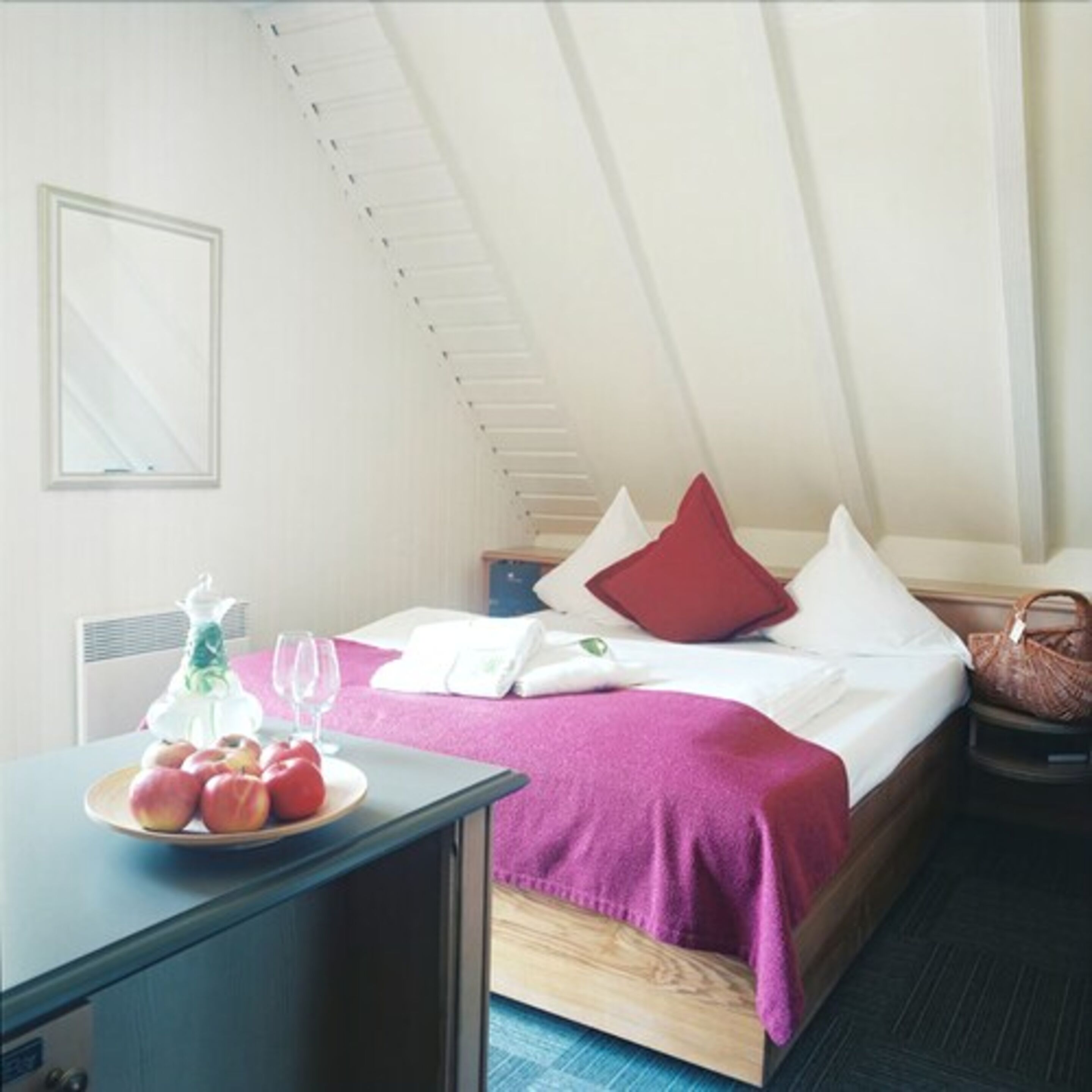 A bedroom with a bed, a bedside table with a fruit bowl, a lamp, and a window.