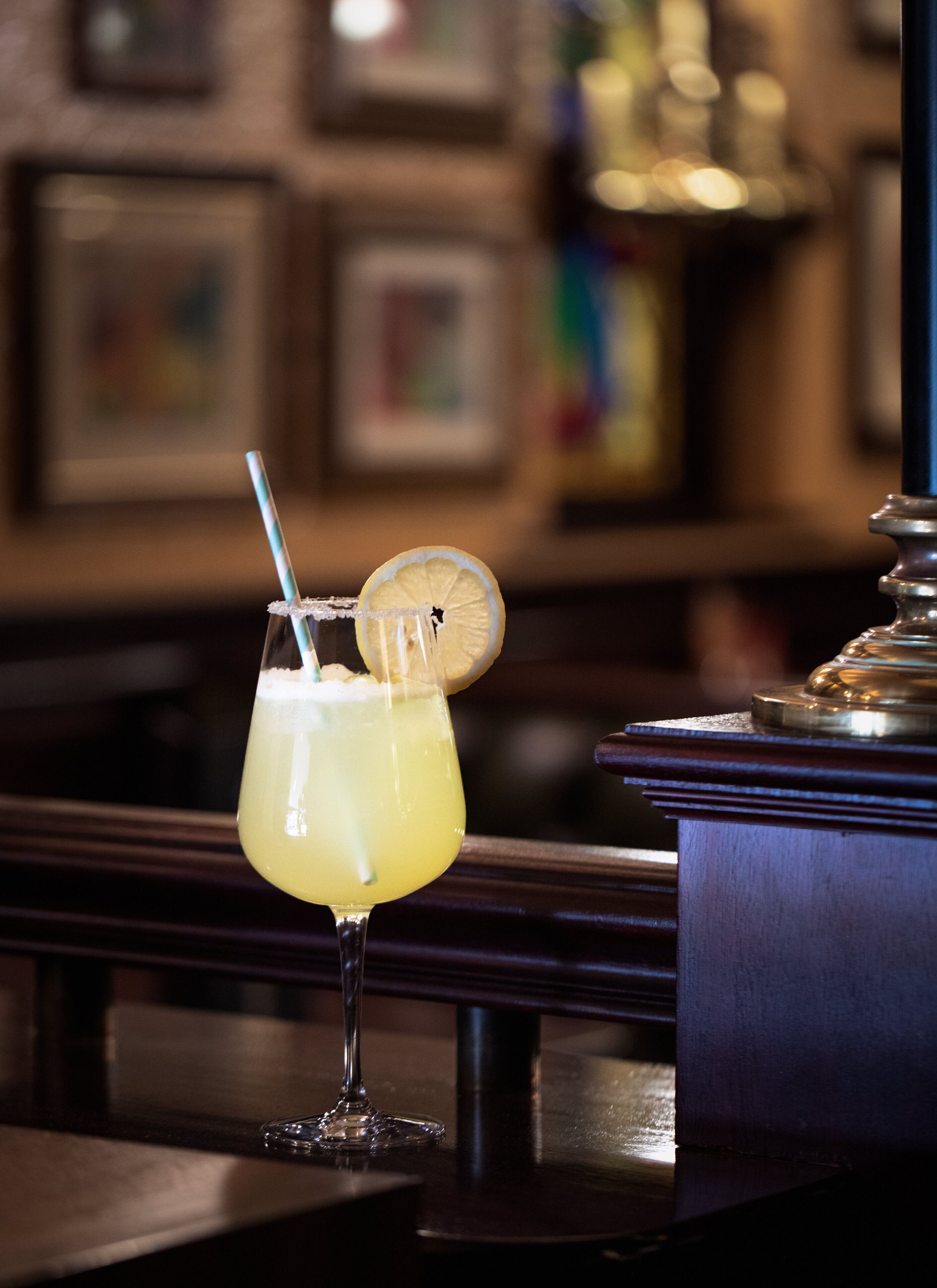 Een cocktailglas met rietje en een citroenpartje op de rand, geplaatst op een barachtergrond met een vage achtergrond van ingelijste foto's en een decoratief object.