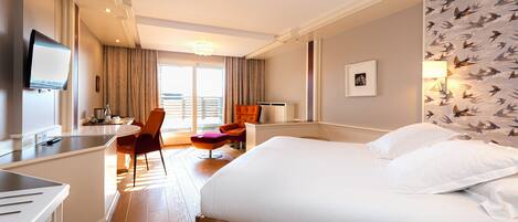 A hotel room with a large bed, a desk with a flat-screen TV, a chair, a small table, and a window with curtains.