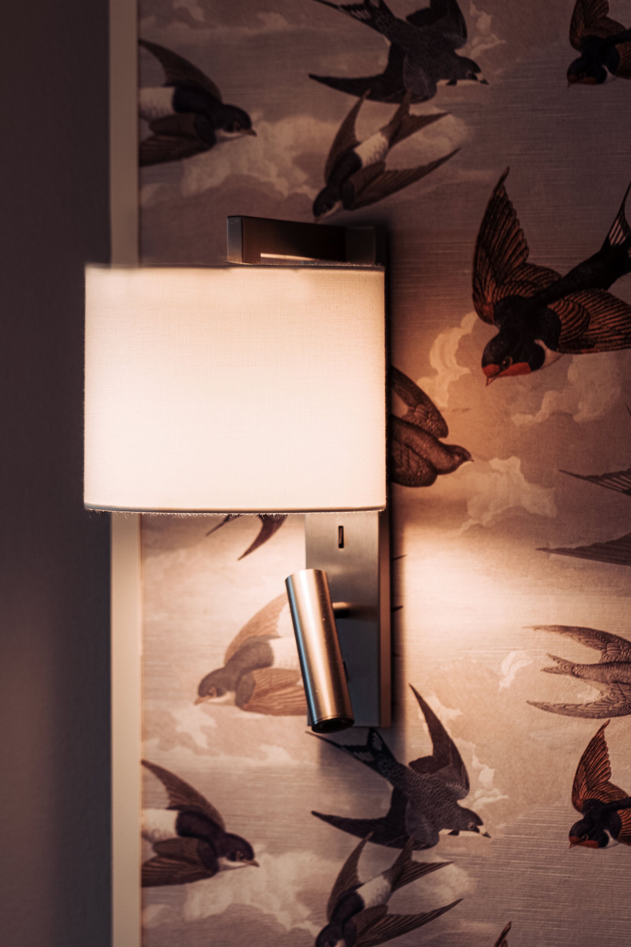 A wall-mounted lamp with a rectangular shade, set against a wallpaper with a bird pattern.