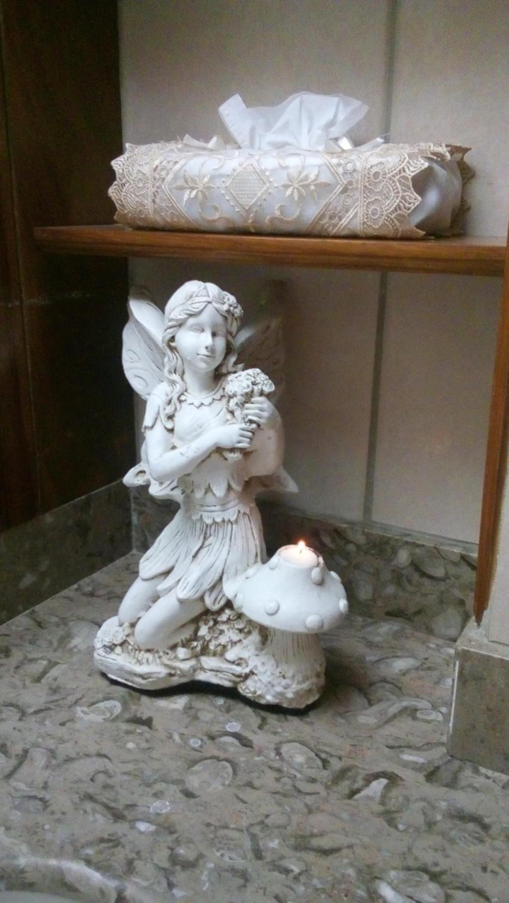 A statue of a fairy holding a flower and a mushroom, placed on a marble floor with a lit candle.