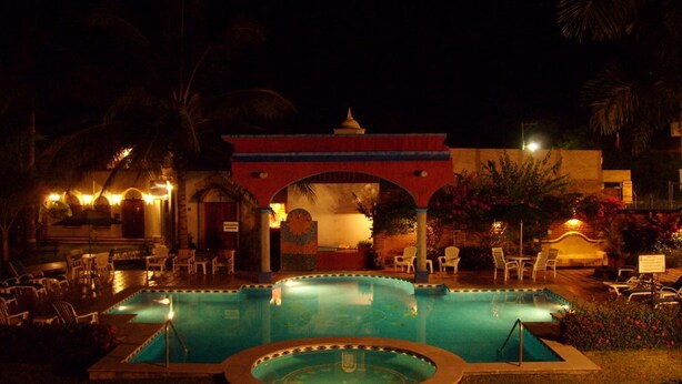 A nighttime pool area with lounge chairs and a building with a red archway. - Hotel Hacienda Flamingos (San Blas)