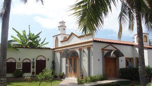 A two-story building with a white facade, red-tiled roof, and a central entrance with a wooden door. There are palm trees and a well-maintained lawn in front. - Hotel Hacienda Flamingos (San Blas)