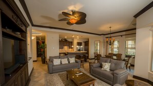 A spacious living room with a large sofa, a coffee table, and a ceiling fan. There is a kitchen area in the background with a dining table and chairs. - Los Suenos Resort Marbella 3D (Jacó)