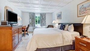 A hotel room with a large bed, a desk with a television, a chair, a desk lamp, and a window with curtains.
