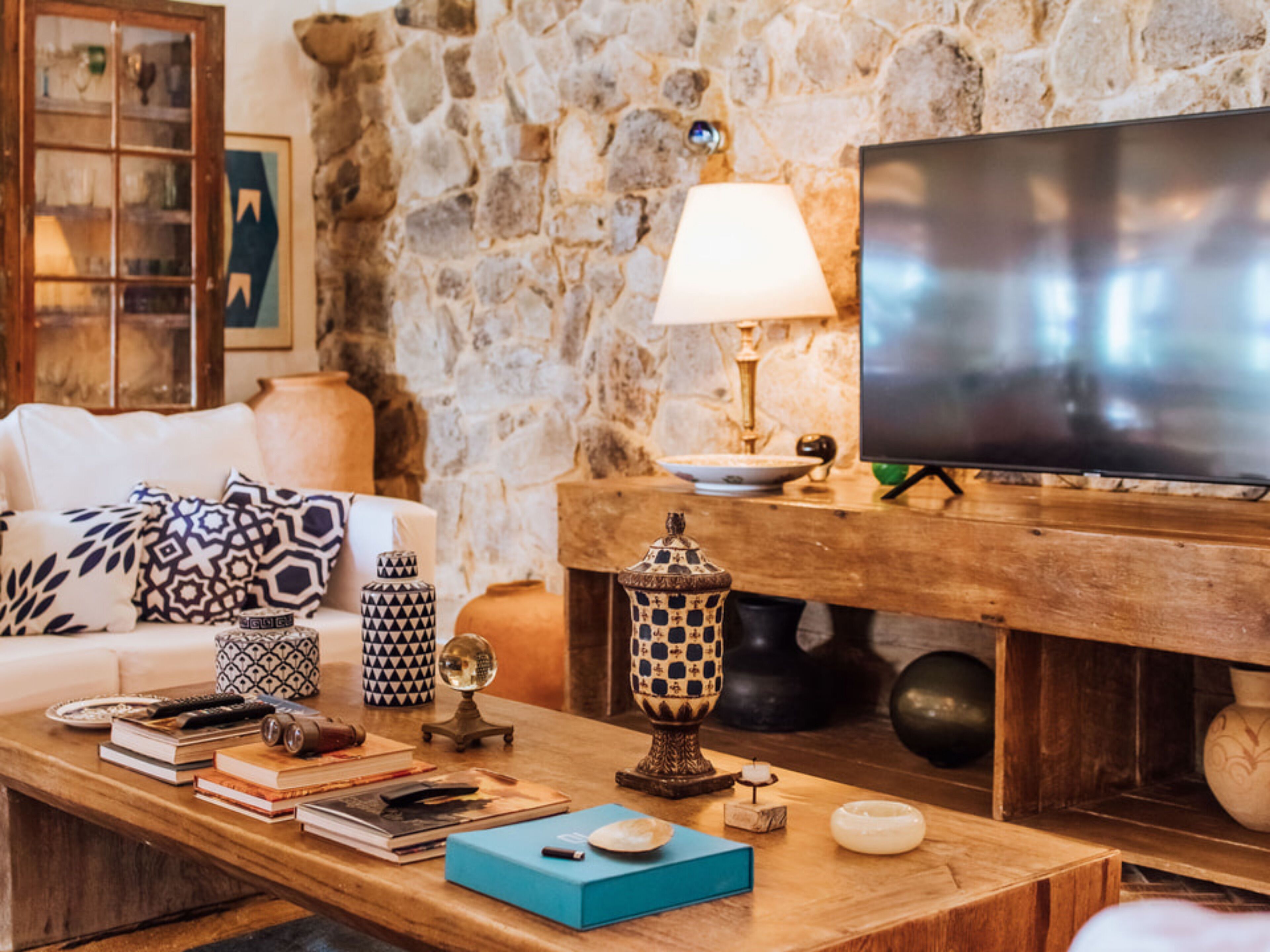 Un salon chaleureux avec un mur en pierre, un téléviseur à écran plat, une table basse en bois agrémentée de livres et d’objets décoratifs, une lampe à motifs et un canapé blanc avec des coussins à motifs.