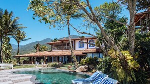 Ein Pool mit Liegestühlen, ein Haus mit Ziegeldach und eine Bergkulisse im Hintergrund.
