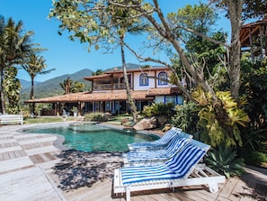 Ein Pool mit Liegestühlen, ein Haus mit Ziegeldach und eine Bergkulisse im Hintergrund.