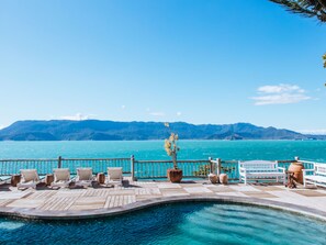 Una terraza de madera con sillones de descanso y piscina, con vistas al mar turquesa y montañas.