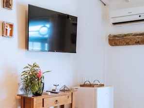 A room with a flat-screen TV mounted on the wall, a wooden desk with a potted plant, a small table with glasses, and a white refrigerator.