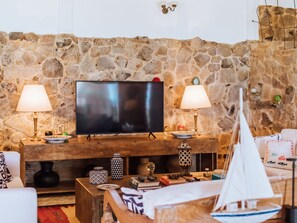 A living room with a stone wall, a flat-screen TV, two lamps, a wooden coffee table, and a sailboat model. - Vilatoro Guest House (Ilhabela)