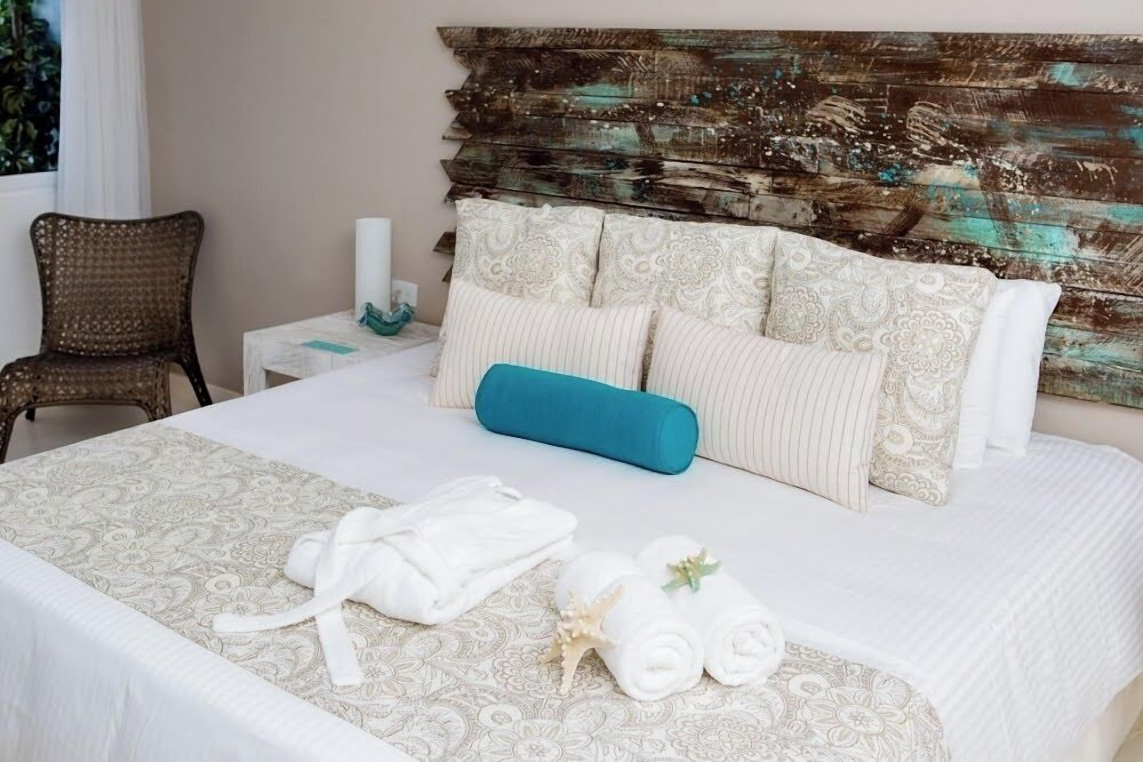 A neatly made bed with a patterned bedspread, a blue pillow, and rolled towels with a seashell.