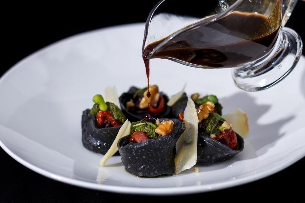 A white plate with a dark-colored food item topped with nuts and greens, and a dark sauce being poured over it.